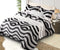 A bedsheet with a black and white zigzag pattern is laid on a bed in a room with a white bed frame, two pillows, and a lamp. The bedsheet is also shown in a flat lay position beside the bed.
